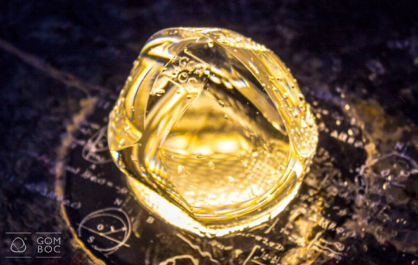 A képen egy fényes Gömböc, sárgás arany színben ragyog. Mögötte fekete alapon árgás matematikai és fizikai képletek.