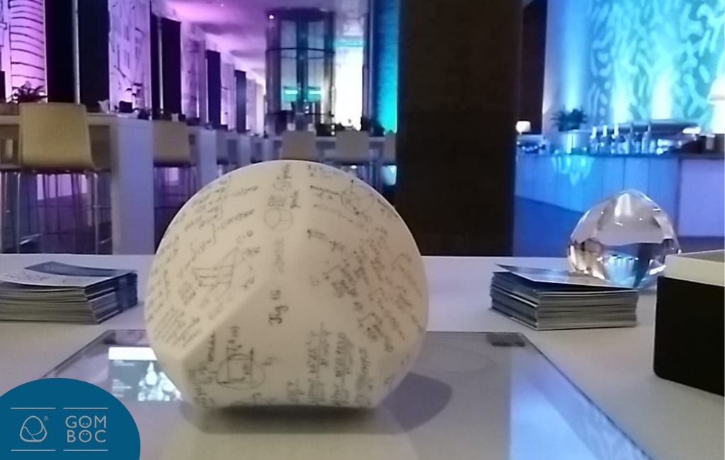 The centerpiece of the photo is a Herend porcelain Gömböc on the table in a large room. There is also a plexiglass Gömböc on the table, along with informational brochures about them.