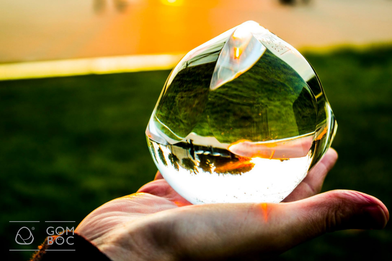 A képen egy kéz látható amint egy plexiüveg Gömböcöt tart. Zöldes, homályos háttér előtt.